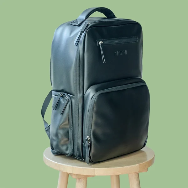 Sleek black diaper backpack with multiple zippered compartments displayed on a wooden stool against a green background